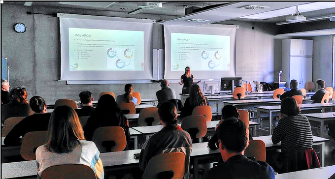 Picture of Saskia Hiltemann presenting the MAdLand tools and resources at the DOMPS plant science seminar. Several attendees can be seen in the foreground, the current slide behind her is entitled “Why ARCs?” and shows a visual representation of the data management lifecycle of research projects.